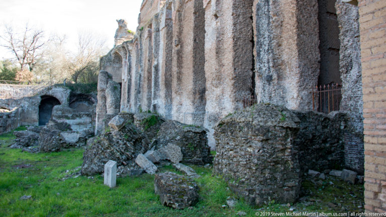 Long weathered Roman masonry wall with repeated vertical masses, rough foundations, grass, and a small arched opening.