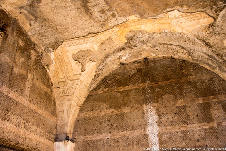 Cream and ochre geometric painted plaster survives in angular bands across a curved masonry vault.