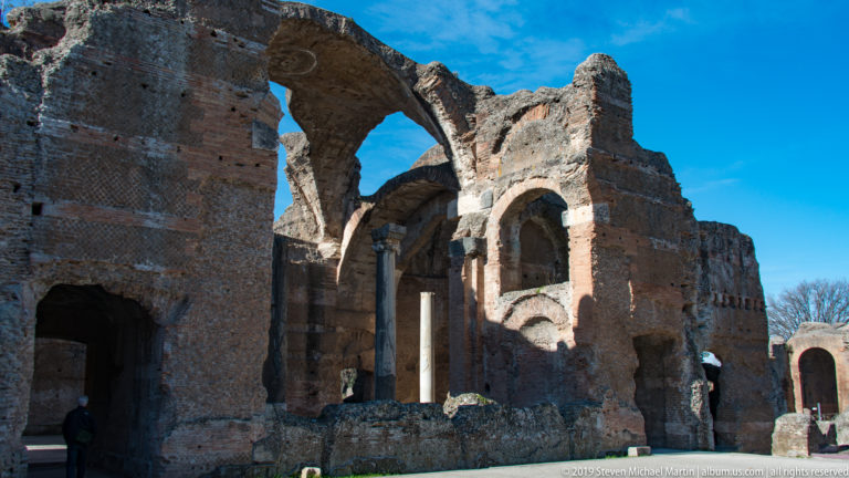 Tall weathered brick ruin with large arches, deep niches, surviving vaults, and slender columns under blue sky.