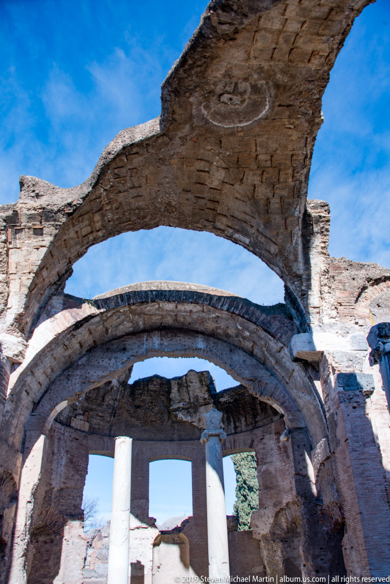 Upward view through intersecting brick arches and curved vaults with a pale column below and blue sky beyond.