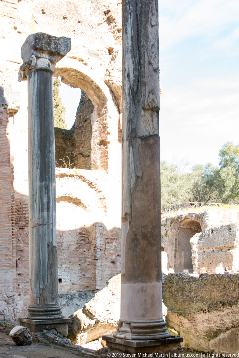 Two stone columns stand beneath large weathered brick arches with sunlit ruins visible through the openings.
