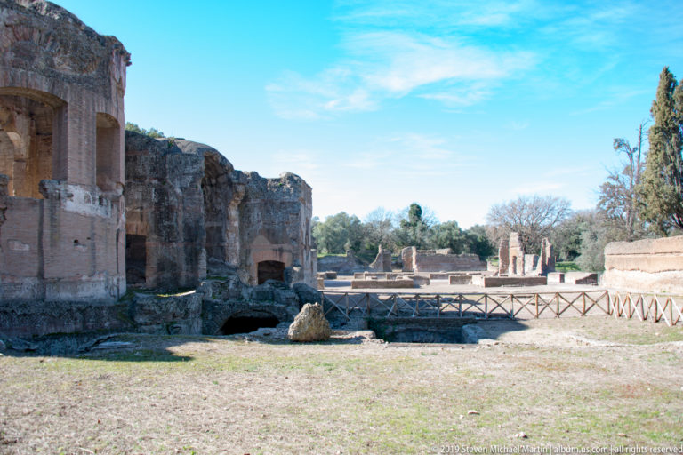 Open grassy archaeological area with low masonry foundations, taller brick ruins, and a wooden railing in the distance.