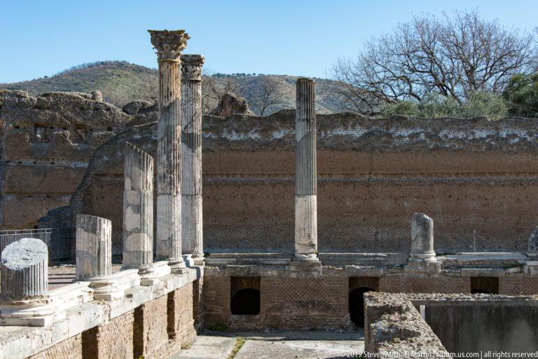 Standing columns line a raised edge above lower brick chambers against a long exposed Roman masonry wall.