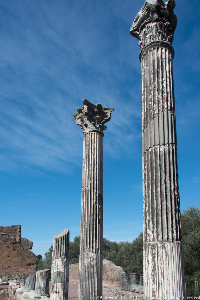 Two tall fluted columns with ornate capitals and a shorter broken shaft rise against a deep blue sky.