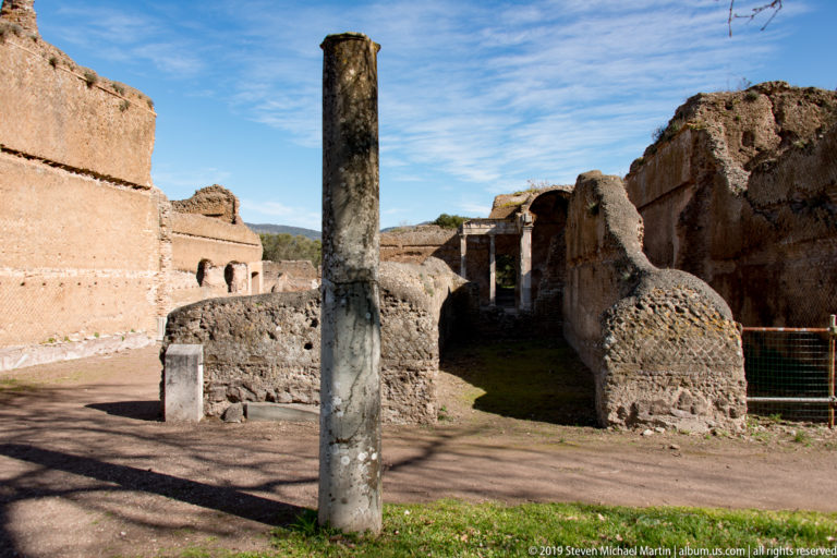 Single standing column before low masonry, small arches, and a narrow passage through weathered brick ruins.