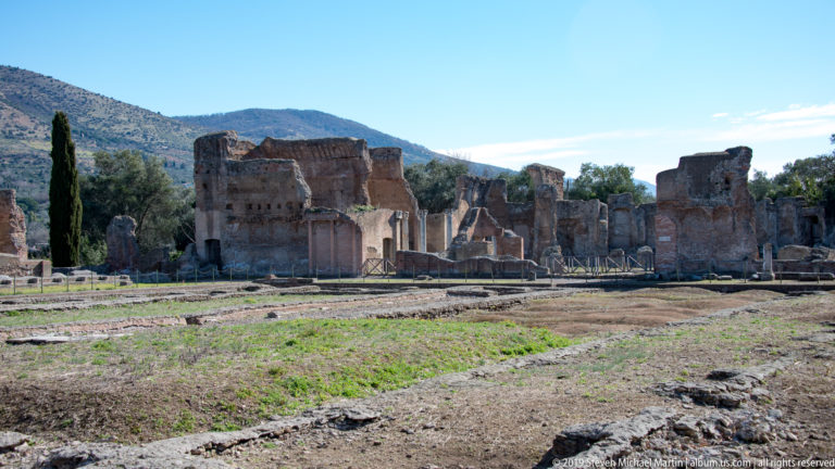 Wide archaeological landscape with low foundations, open ground, and a line of tall brick ruins beneath distant hills.