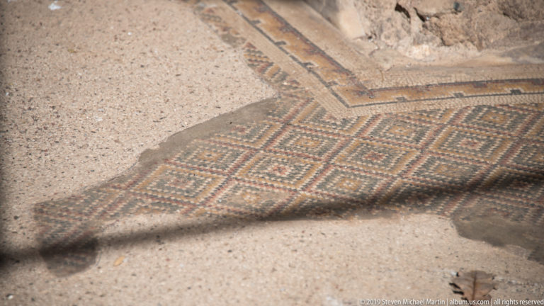 Fragmentary geometric mosaic pavement with repeated angular patterns and a narrow decorative border beside exposed substrate.