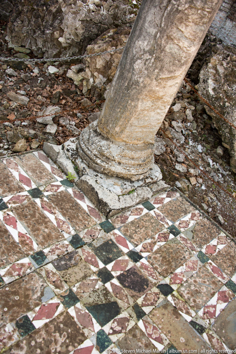 Column base beside a geometric multicolored stone pavement of red, white, green, and dark triangular pieces.