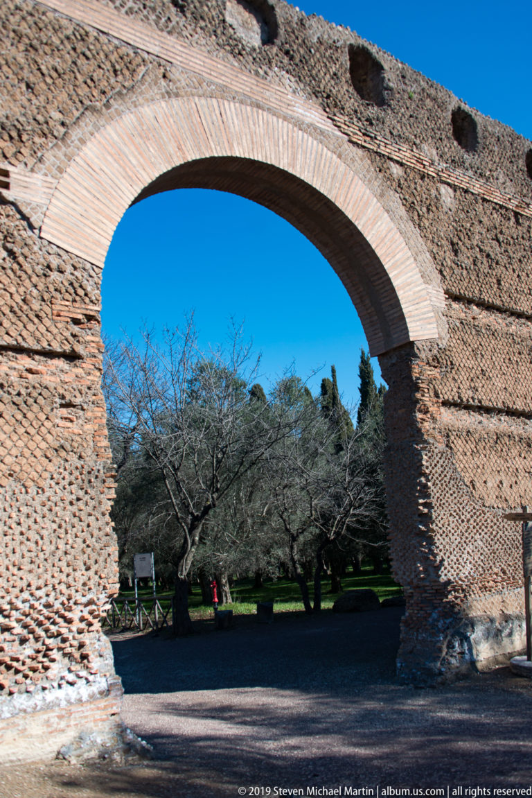 Large round brick arch in a thick weathered Roman wall, framing leafless trees and the landscaped grounds beyond.