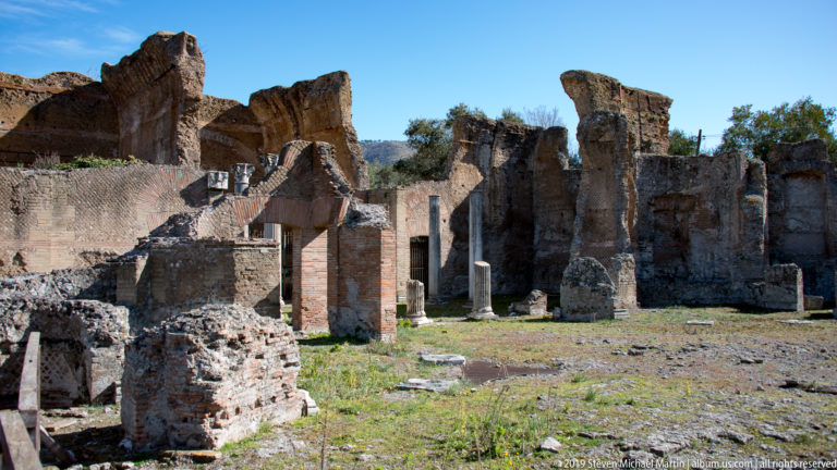 Open archaeological court with tall broken brick walls, doorways, low foundations, and scattered column shafts.