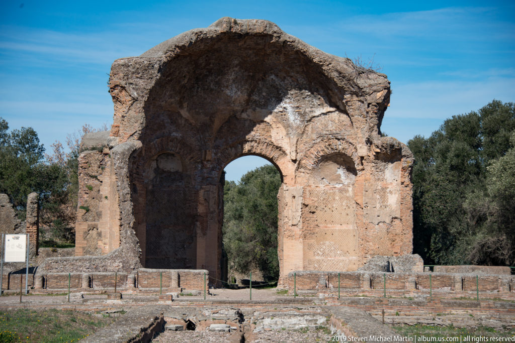 Large weathered Roman arch with a central opening standing against blue sky and trees at Hadrian’s Villa.