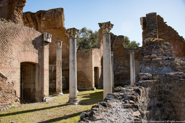 Three tall fluted columns stand among sunlit brick walls, arches, and rough masonry ruins at Hadrian’s Villa.