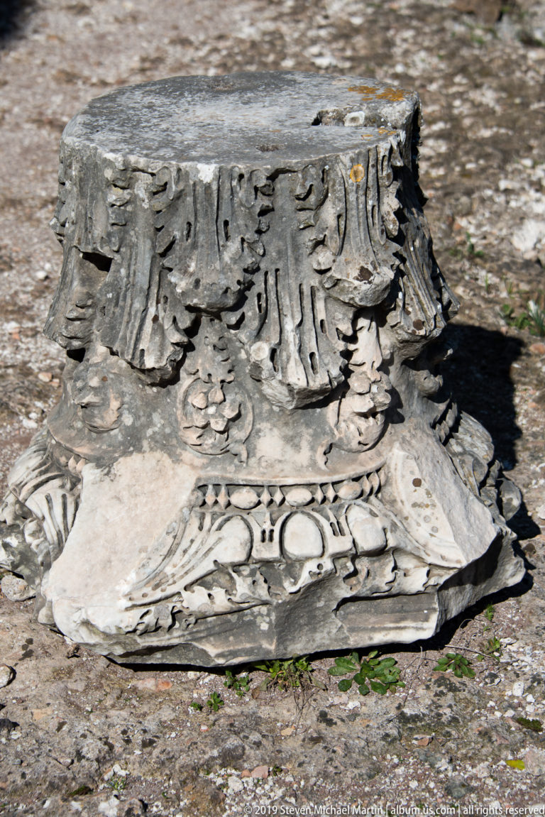 Broken carved stone fragment with layered leaf and scroll ornament resting on the archaeological ground.