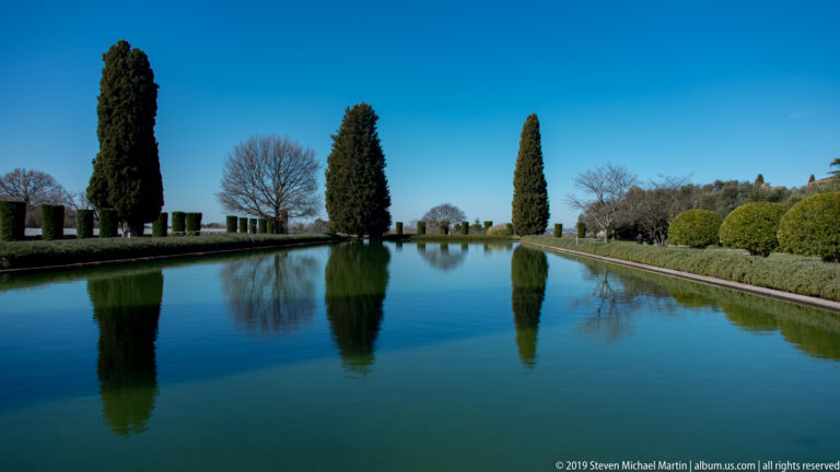 Long rectangular reflecting pool bordered by clipped hedges and tall cypress trees under a clear blue sky.