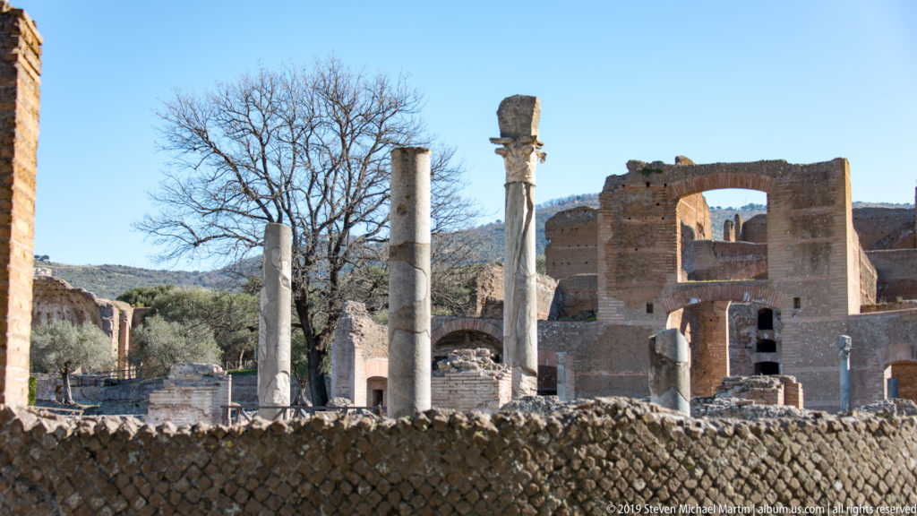 Standing columns and multistory brick Roman ruins with large openings behind a low patterned masonry wall at Villa Adriana.