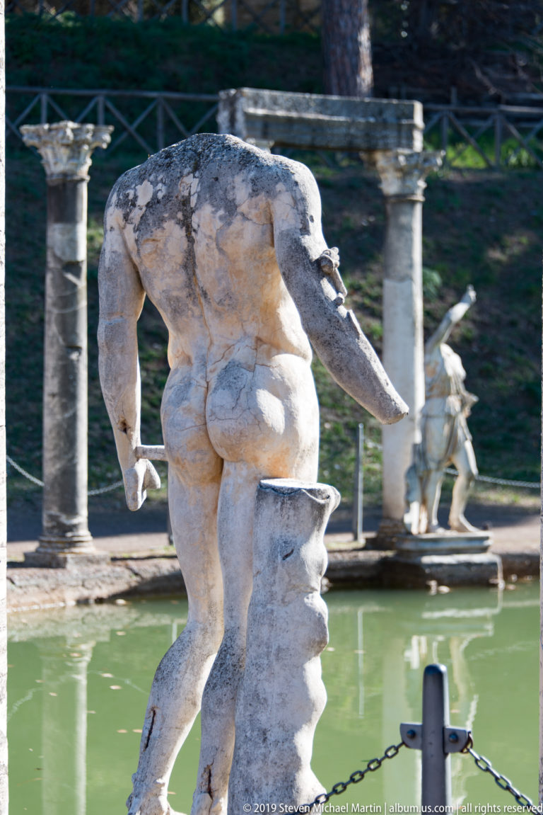 Headless male stone statue leaning forward above the Canopus, seen from behind with columns and another sculpture across the water.