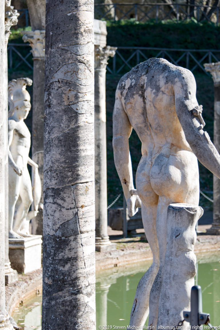 Rear view of a headless weathered male statue leaning forward beside a column and the green Canopus pool.