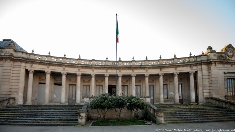 Curved pale-stone colonnade reached by broad steps, with balustrades and an Italian flag rising above the roofline.