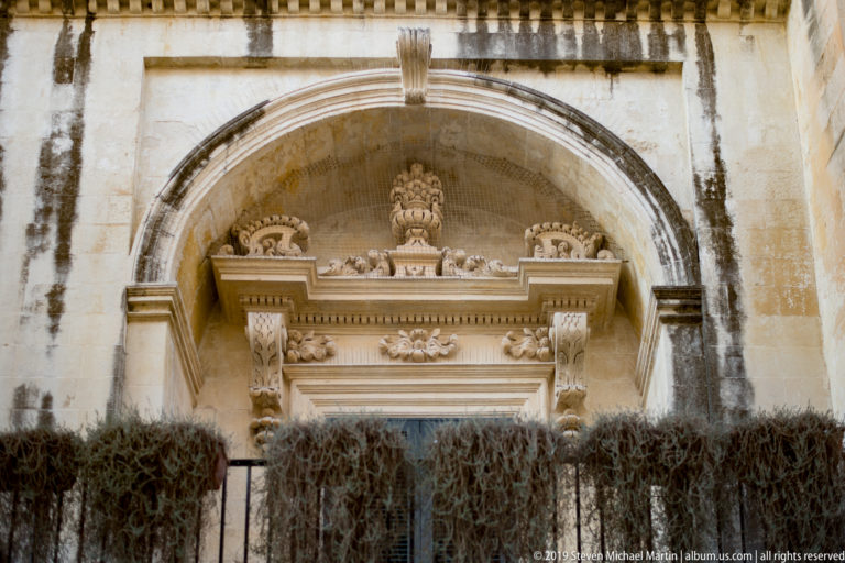 Large arched stone surround framing an ornate portal with crest, projecting cornice and carved consoles on a weathered wall.
