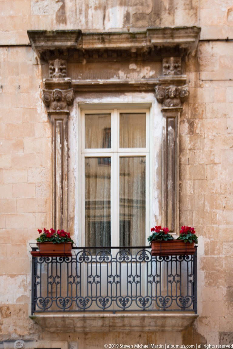 White-framed window with red flower boxes and black wrought-iron balcony set in a weathered limestone façade.