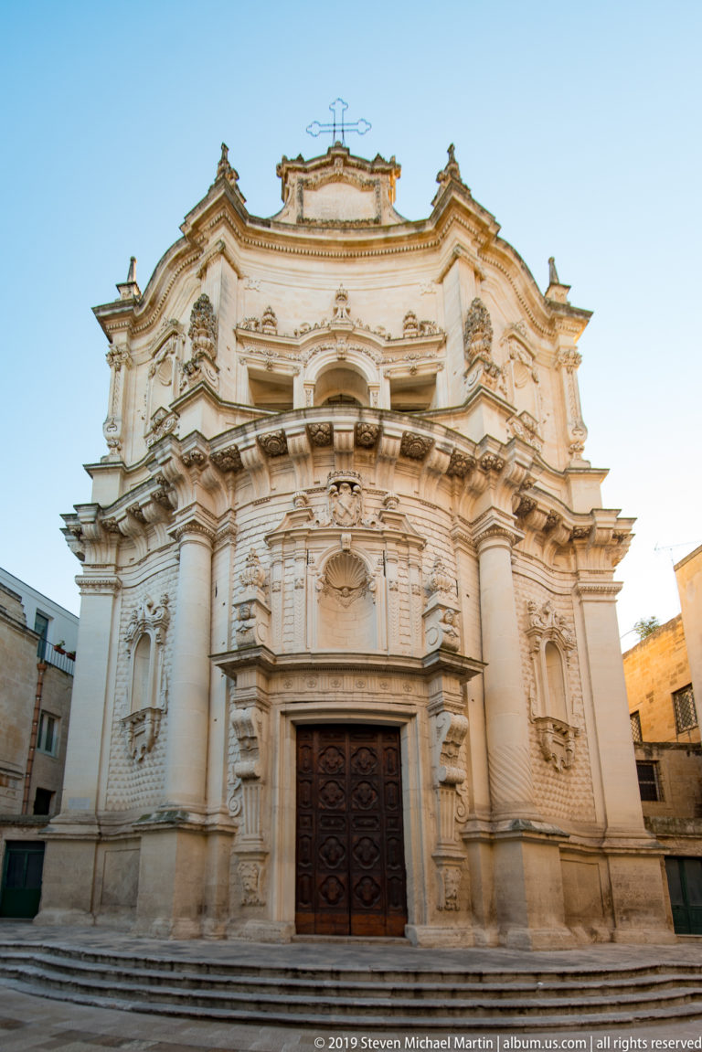 Frontal view of a pale Baroque church with paired columns, niches, layered cornices and a large dark wooden doorway.