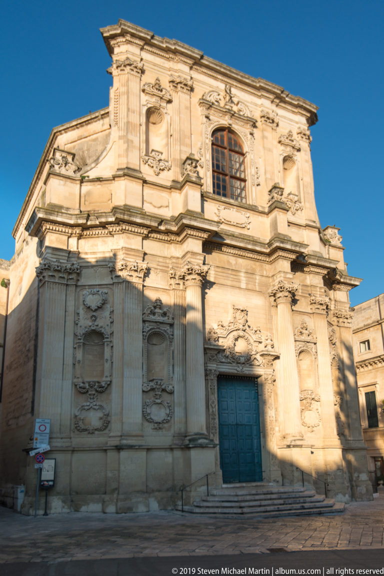 Angled view of a pale Baroque church façade with giant pilasters, carved panels, upper window and blue central door.