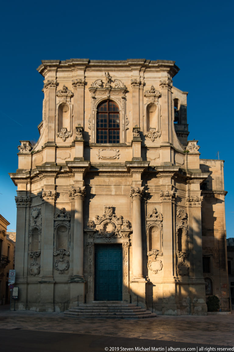 Frontal Baroque church façade with blue door, pilasters and carved niches partly crossed by strong late-day shadow.