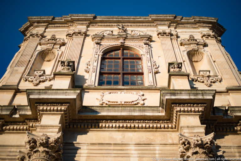 Upward view of ornate pale-stone façade with large window, pilasters, carved reliefs and projecting cornices against blue sky.