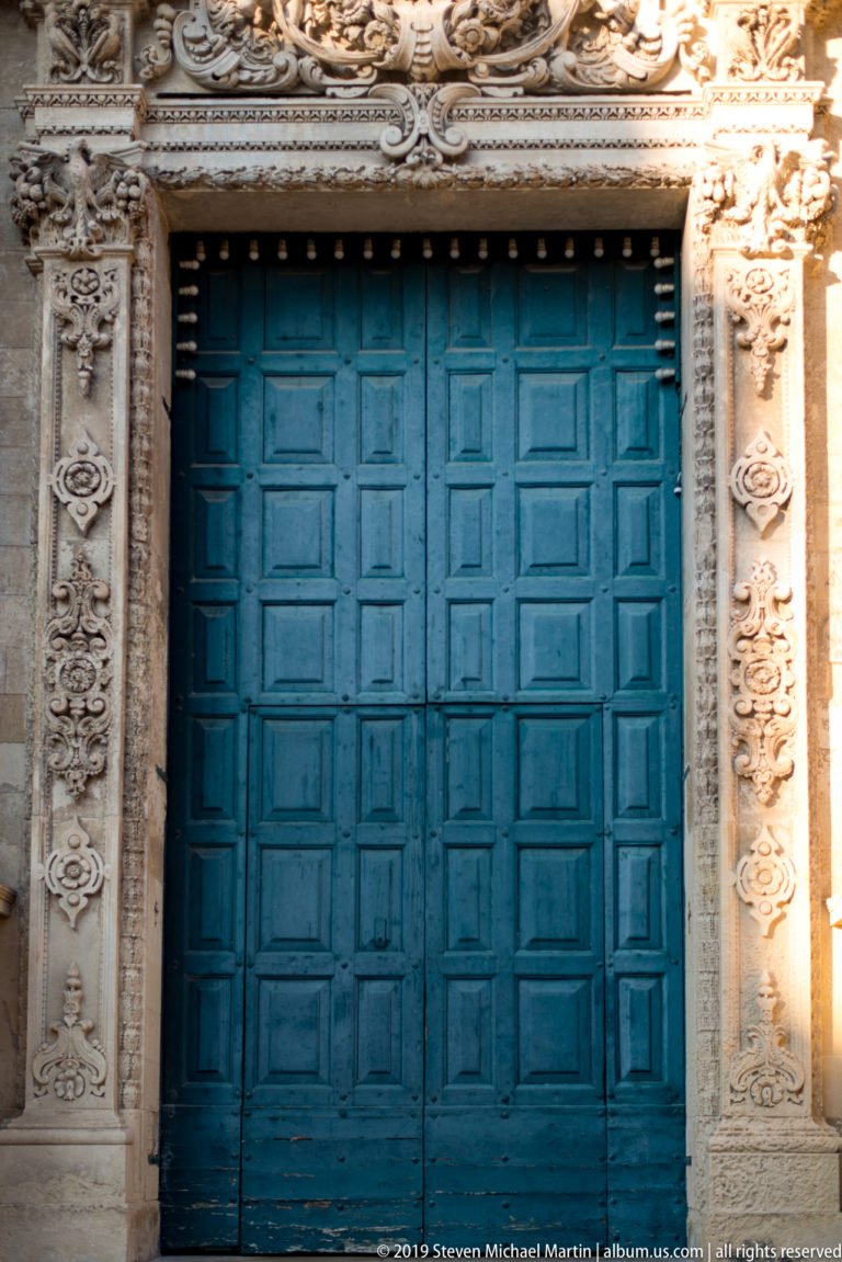 Large blue paneled double doors framed by pale carved stone with floral and scroll ornament.