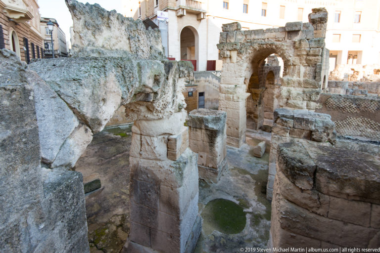 Excavated ancient stone walls, broken arches and massive masonry blocks with modern buildings visible beyond.