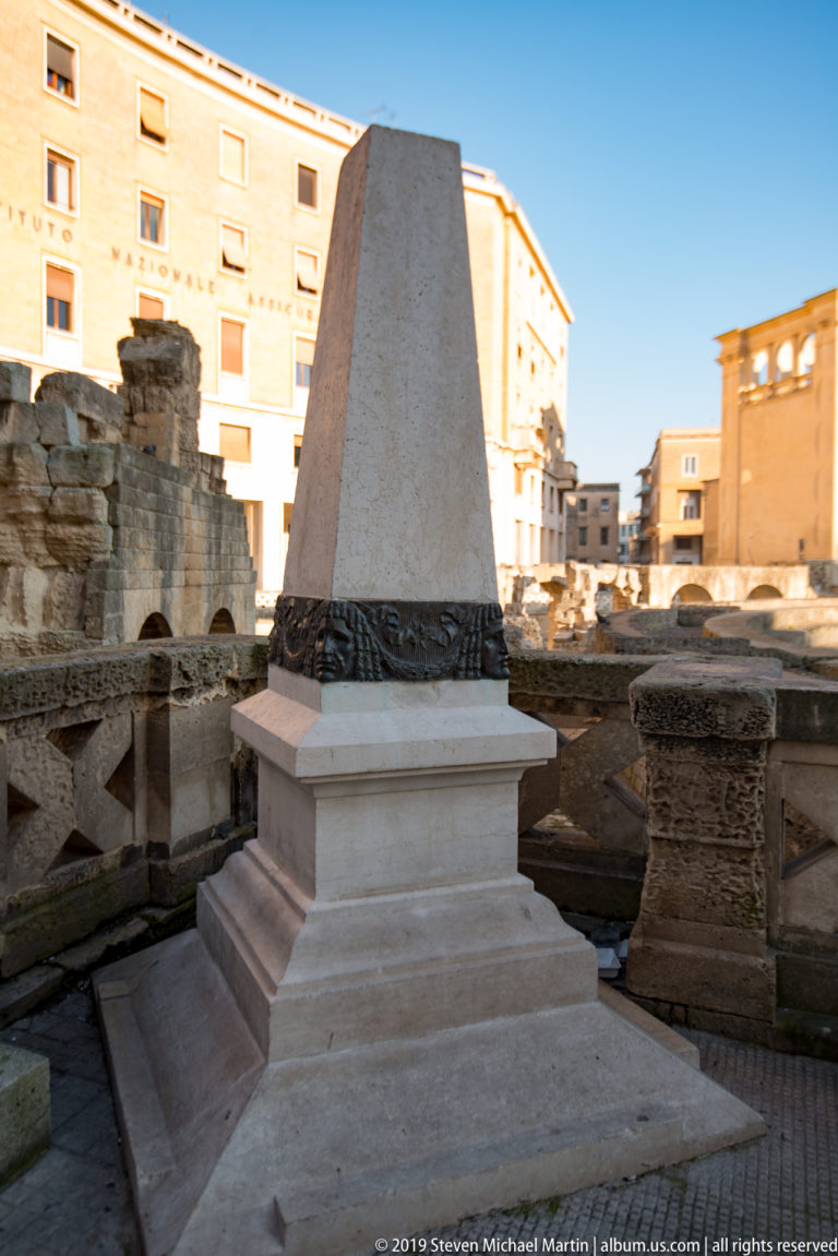 Tapered pale monument with a dark decorative band standing among excavated stone walls at an archaeological site.