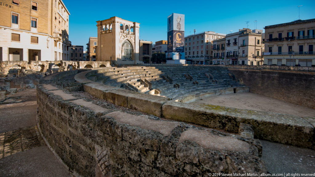 Roman Amphitheatre of Lecce with curved stone seating below the surrounding square and city buildings.