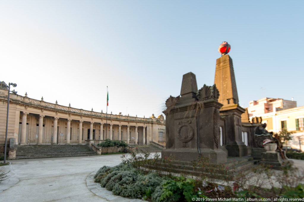 Stone memorial with tall obelisk in a landscaped square, backed by a curved colonnade and Italian flag.