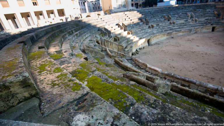 Close view of curved, weathered stone seating tiers in Lecce’s Roman Amphitheatre, with patches of moss and exposed passages.