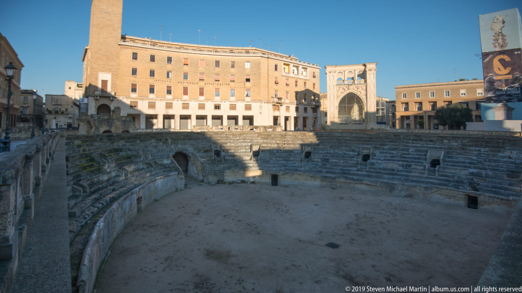 Wide view across the Roman Amphitheatre’s arena toward curved seating and multi-story buildings around the square.