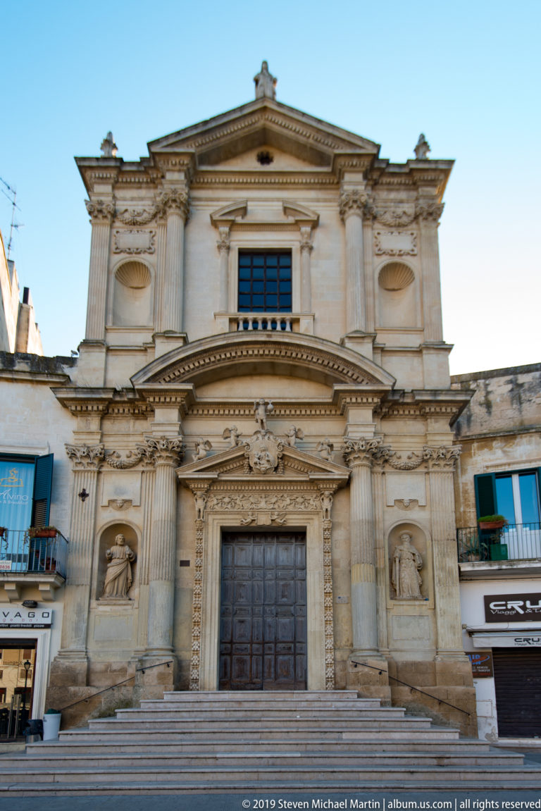 Symmetrical pale-stone church façade above broad steps, with pilasters, triangular pediments, niches and a dark wooden door.