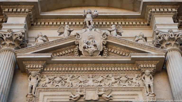 Ornate limestone pediment with putti, heraldic motifs, garlands and dense Baroque relief carving between columns.