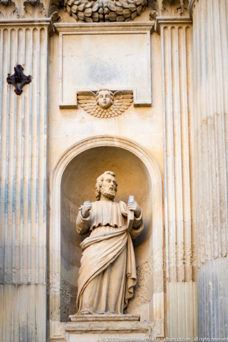 Stone statue of a robed saint in a shallow niche, with a small winged head carved above the arch.