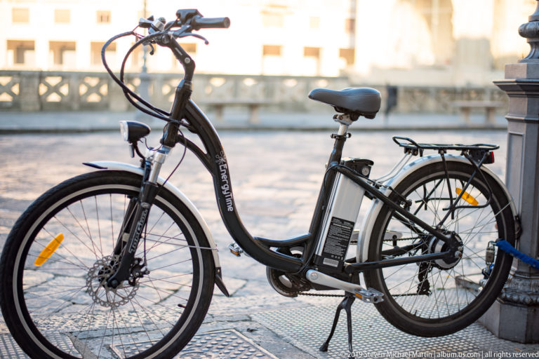 Black step-through electric bicycle with battery and rear rack parked beside pale stone balustrade in Lecce.