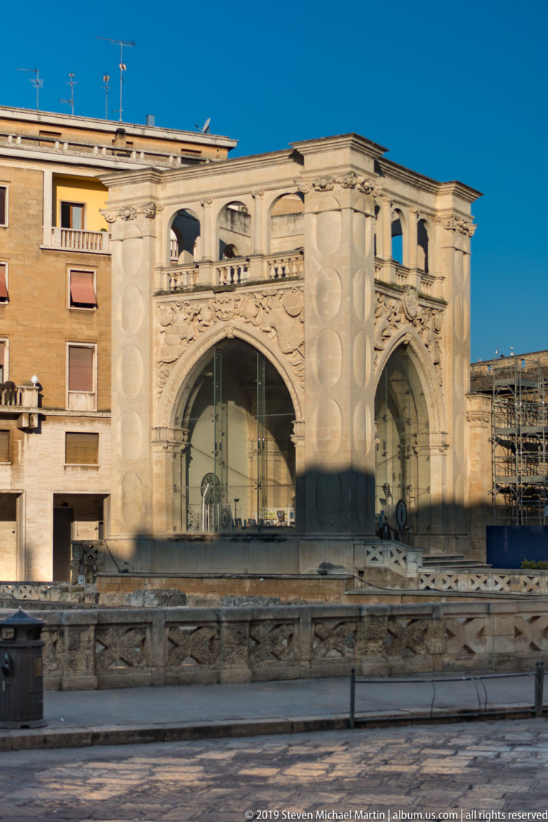 Large pale structure with two broad glazed arches, circular wall panels and balustraded upper level beyond archaeological stonework.