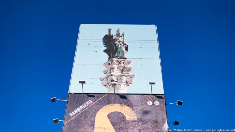 Vertical outdoor display showing a photographed sculptural monument above a large graphic letter, with spotlights projecting from both sides.