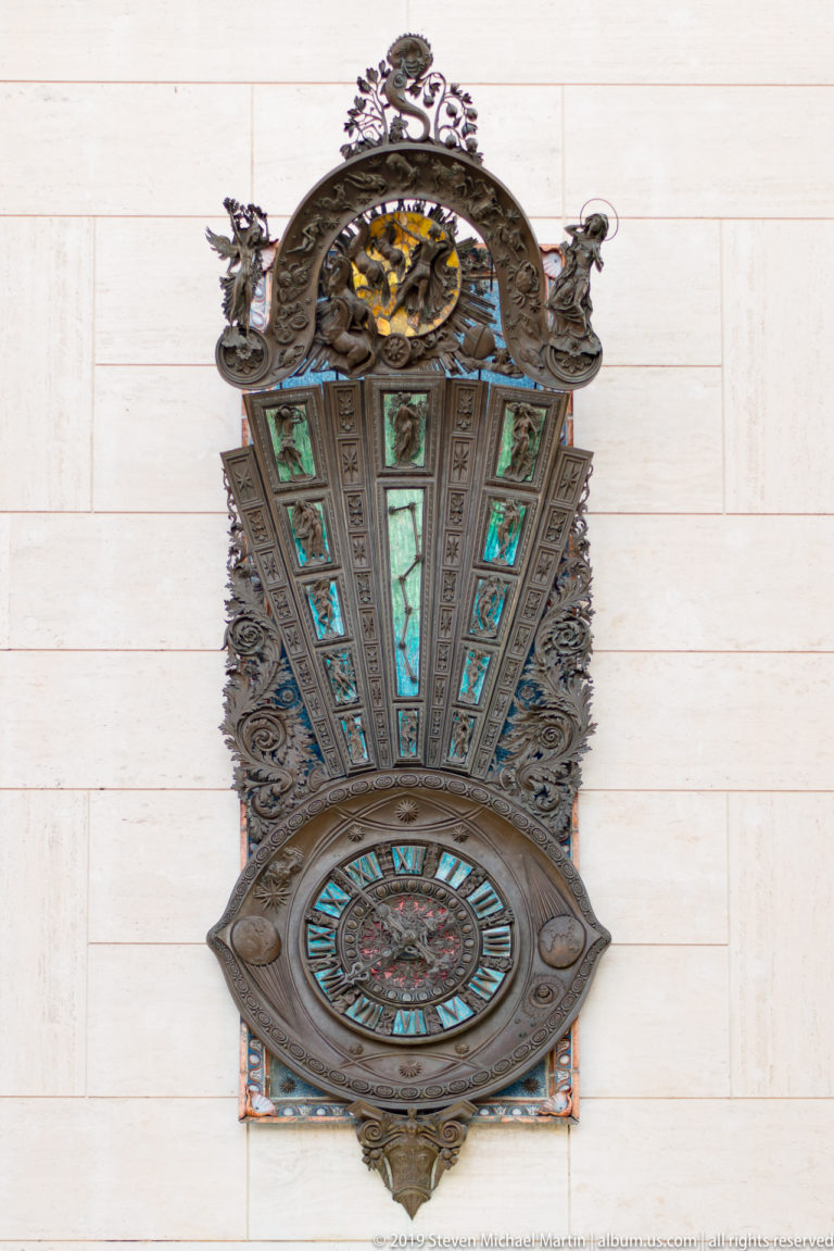 Elaborate projecting wall clock with circular dial, ornate dark metal frame and turquoise-green decorative panels.