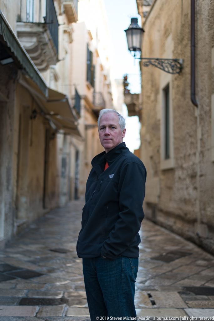Man in dark coat standing on a narrow stone-paved Lecce lane, with historic buildings and a street lamp behind him.