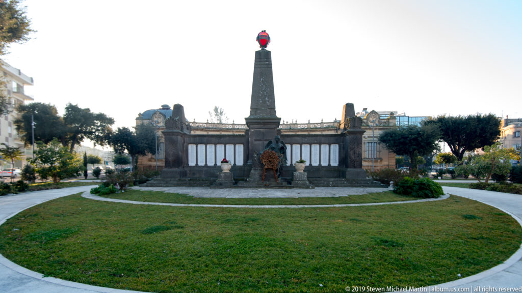 Central obelisk memorial beyond a circular lawn, surrounded by formal plantings and low urban buildings.