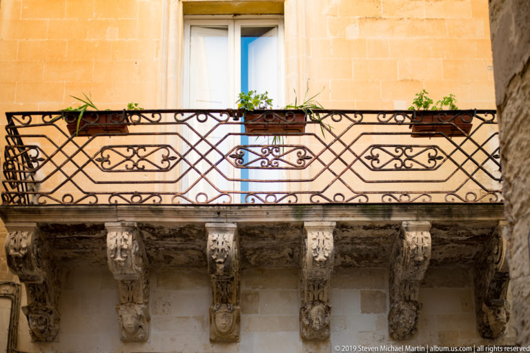 Wrought-iron balcony with potted plants above a row of carved stone corbels on a pale limestone façade.