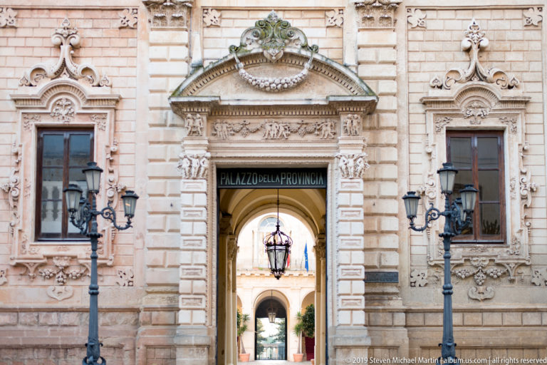 Ornate Palazzo della Provincia entrance with carved pediments, tall windows, lamps and an arched passage into a courtyard.
