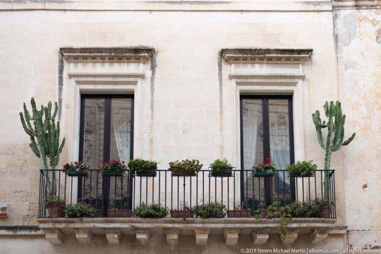 Iron balcony with two tall windows, red flower boxes and large cactus plants on a pale stone façade.
