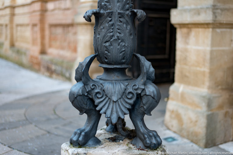 Dark cast-metal street-lamp base with three paw-shaped feet, scrolling ornament and a leaf-decorated shaft.