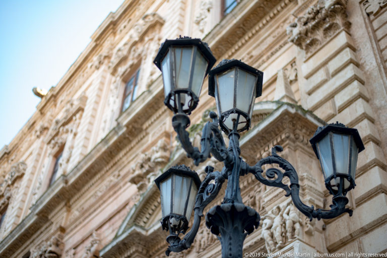 Four-lantern ornamental street light with scrolling cast-metal arms against a richly carved pale stone façade.