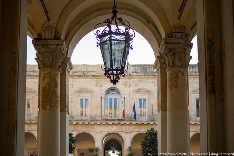 Large hanging lantern beneath an arch between stone columns, looking toward a sunlit courtyard façade with flags.